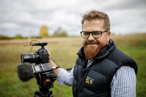 Smiling Phil standing in a green field holdinga DSLR camera